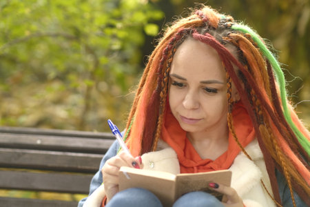 Young woman student with long hair in casual clothes sitting on wooden bench and writing notes in notebook.の写真素材