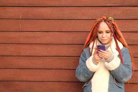 Woman with orange dreadlocks and in a warm denim jacket using smartphone on red wooden background.