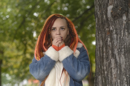 Young frozen woman with dreadlocks standing near tree in cold weather. Female with colorful...