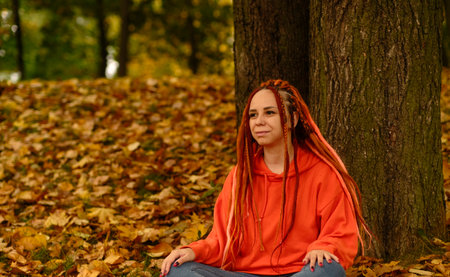 Young woman with dreadlocks sitting near tree in golden autumn. Vivid female with colorful hairstyle...