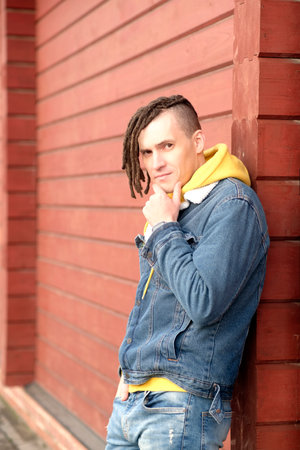 Portrait of young positive man with dreadlocks standing, leaning on wall, on city street