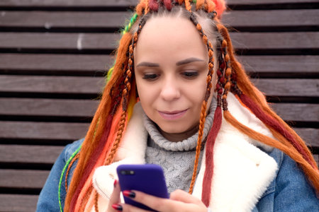 Young woman with dreadlocks in warm clothes browsing mobile phone, sitting on wooden sunbed in city park. Carefree female with colorful hairstyle using smartphone on walk in cloudy weatherの写真素材