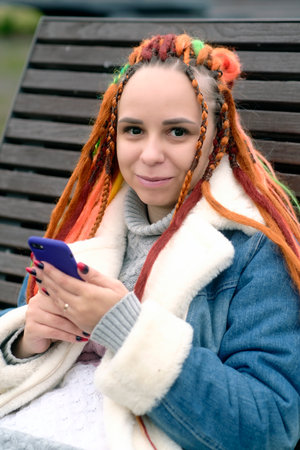 Young woman with dreadlocks in warm clothes browsing mobile phone, sitting on wooden sunbed in city park. Carefree female with colorful hairstyle using smartphone on walk in cloudy weatherの写真素材