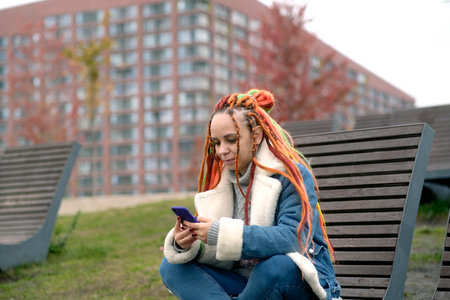 Young woman with dreadlocks in warm clothes browsing mobile phone, sitting on wooden sunbed in city park. Carefree female with colorful hairstyle using smartphone on walk in cloudy weatherの写真素材