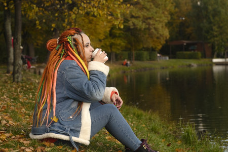 Young woman with dreadlocks in warm clothes sitting on the shore of the lake drinking coffee in park.の写真素材