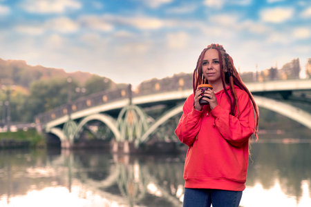Young woman with dreadlocks holding cup of coffee, standing near river, lake on background of big bridge in city parkの写真素材