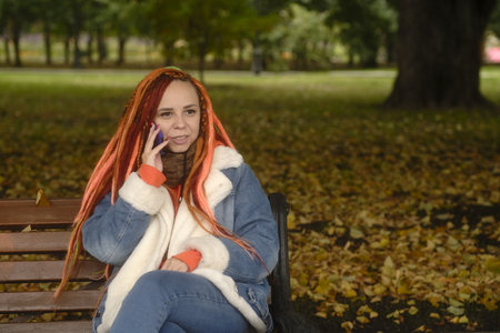 Attractive young woman in warm clothes sitting on bench and talking on mobile phone in park.の写真素材