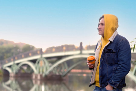 Young man with dreadlocks holding cup of coffee, standing near river, lake on background of big...