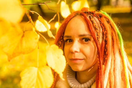 Portrait of young positive woman with ginger dreadlocks standing among branches of tree with yellow...
