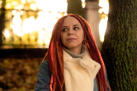 Portrait of young positive woman with ginger dreadlocks looking around, standing near tree trunkの写真素材