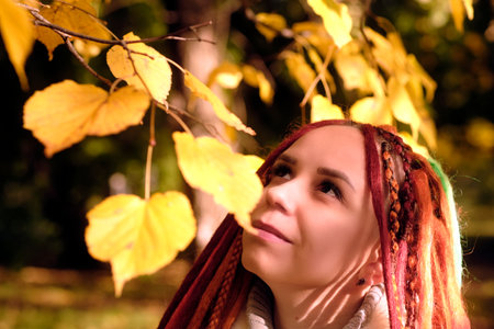 Portrait of young positive woman with ginger dreadlocks standing among branches of tree with yellow leavesの写真素材