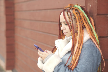 Woman with orange dreadlocks and in a warm denim jacket using smartphone on red wooden background.の写真素材