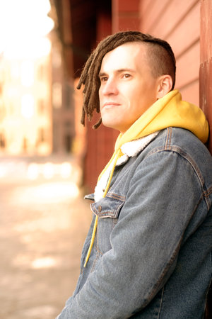 Portrait of young positive man with dreadlocks standing, leaning on wall, on city street