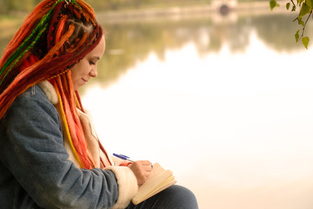 Young creative woman with dreadlocks drawing, writing in notebook, sitting on stumps near lake in...