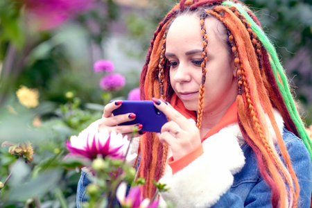 Woman taking pictures on smartphone in park. Positive young female hipster with long dreadlocks in...
