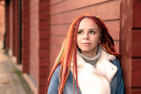 Portrait of positive young woman with dreadlocks, standing leaning on wall, on city street. Pretty...