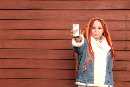Positive young woman with dreadlocks looking at camera, smiling, showing mobile phone with screen...