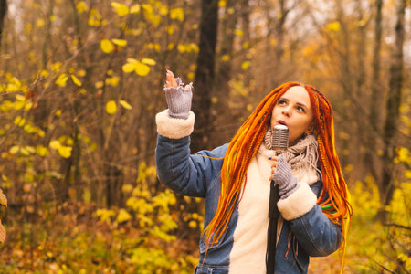 Young woman with bright hairstyle with retro microphone in autumn forest. Portrait of female singer with dreadlocks singing into microphone in natureの写真素材