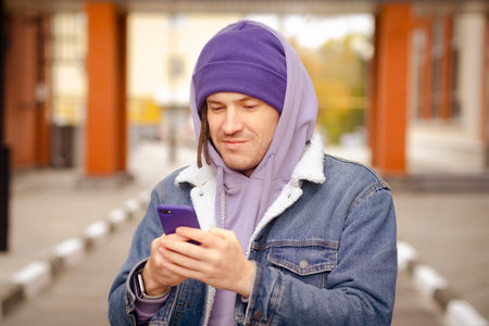 Positive young male in warm clothes smiling and browsing mobile phone while standing on city street.の写真素材