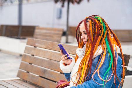 Young woman with bright dreadlocks with mobile phone on walk in park. Portrait of positive female in warm clothes browsing smartphoneの写真素材