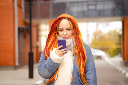 Young woman with bright hairstyle in warm clothes browsing mobile phone, standing on city streetの写真素材