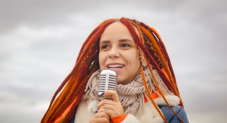 Young woman with dreadlocks singing into microphone on background of cloudy sky. Female singer...