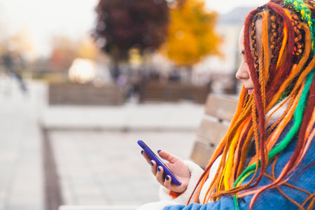 Young woman with bright dreadlocks with mobile phone on walk in park. Portrait of positive female in warm clothes browsing smartphoneの写真素材
