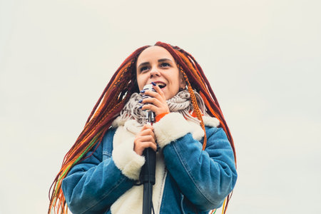 Young woman with dreadlocks singing into microphone on background of cloudy sky. Female singer...
