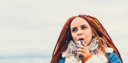 Young woman with dreadlocks singing into microphone on background of cloudy sky. Female singer enjoying singing in natureの写真素材