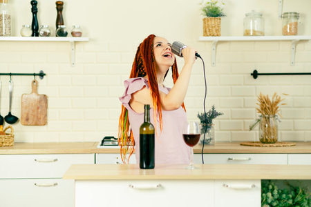 Young woman with bright hairstyle with retro microphone in kitchen. Portrait of female singer with dreadlocks singing into microphone at homeの写真素材