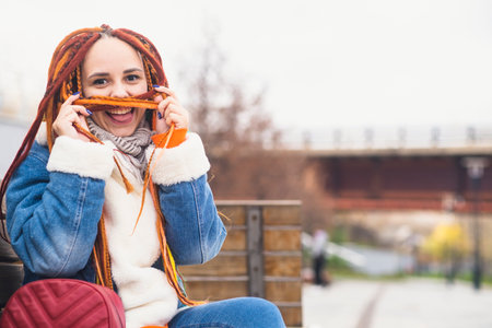 Young playful woman with bright hairstyle in warm clothes on walk in park. Positive female making mustache from dreadlocks, laughing, having funの写真素材