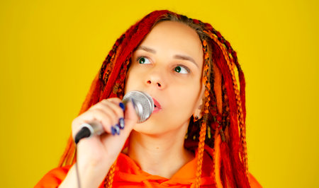 Young woman with bright dreadlocks singing into microphone on yellow background. Emotional female singer with mic in studio. close up.の写真素材