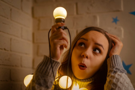 Young thoughtful woman wrapped in garland of glowing bulbs scratching head, raising lightbulb up,...