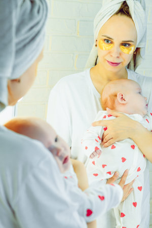 Young mother take care of her skin with under eye patches holding her newborn baby in her arms in...
