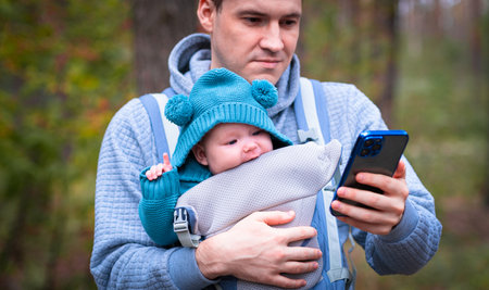 Father and newborn baby in a sling in the autumn forest, man with cellphone. Happy family.の写真素材