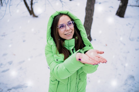 Portrait of a beautiful girl in a green jacket and glasses in the winter forest catching snowflakes.