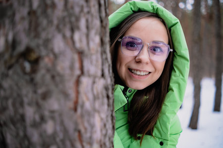 A beautiful woman in a green jacket and wearing glasses hides behind a tree in a winter forestの写真素材