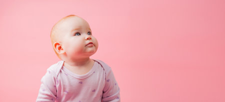 Adorable baby girl sitting on pink background with copy space and looking away.