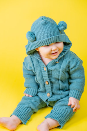 Portrait of a cute baby in a warm knitted jumpsuit with a hood on a yellow background.
