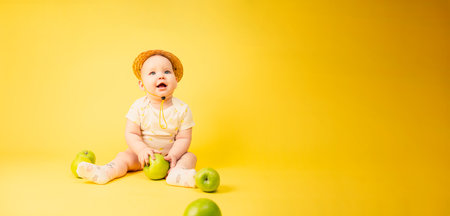Beautiful baby girl in straw hat with green apples sitting in studio on yellow backgroundの写真素材