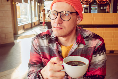 A man with glasses and a orange hat enjoying a cup of coffee sitting in a coffee shop.