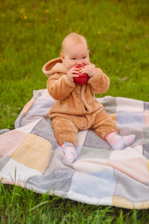 Cute baby girl sitting on plaid and eating apple on the green lawn.