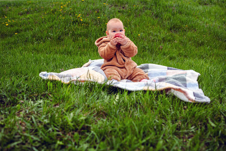 Cute baby girl sitting on plaid and eating apple on the green lawn.