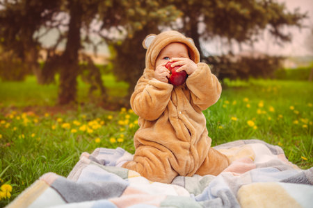 Cute baby girl in plush suit with red apple in hands sitting on blanket in green grassの写真素材