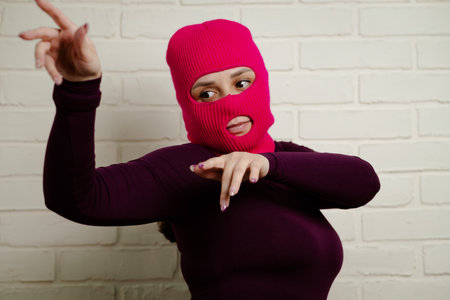 A woman wearing a bright pink balaclava dances energetically, striking unique poses with her hands...