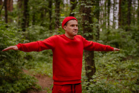 A professional sportsman in a red sweater is standing in the woods, doing shoulder and arm stretches as part of his warm-up routine before a workout sessionの写真素材