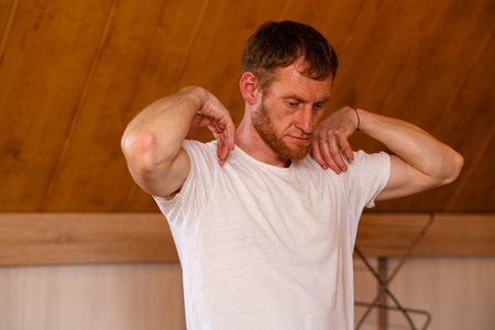 A professional sportsman in a white shirt is standing, holding his hands on his head as he performs shoulder and arm stretches. Warming up before a workout or training sessionの写真素材