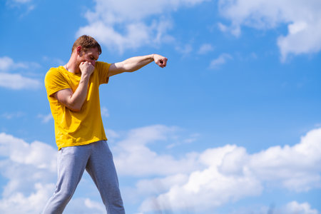 A man wearing a yellow T-shirt is practicing his boxing techniques outdoors against a backdrop of...
