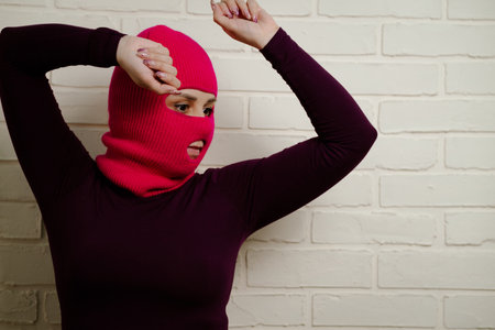 A woman wearing a bright pink balaclava dances energetically, striking unique poses with her hands against a clean, white brick wall background.の写真素材