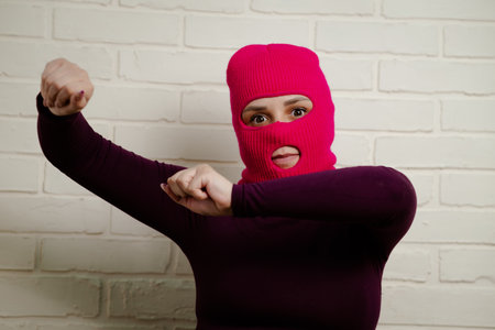 A woman wearing a bright pink balaclava dances energetically, striking unique poses with her hands against a clean, white brick wall background.の写真素材
